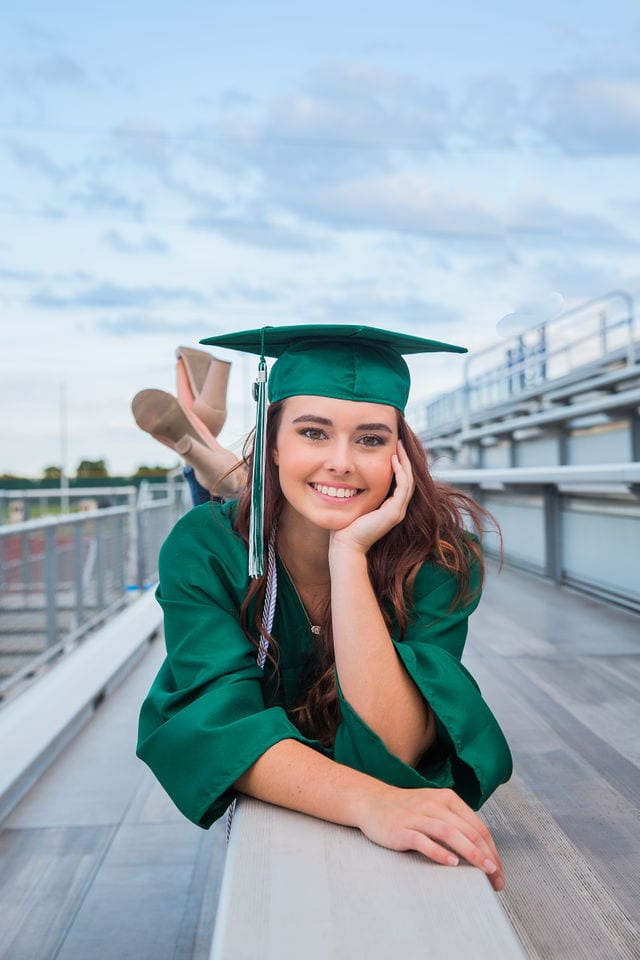 Faith Senior session in Marion cap and gown laying on bleachers